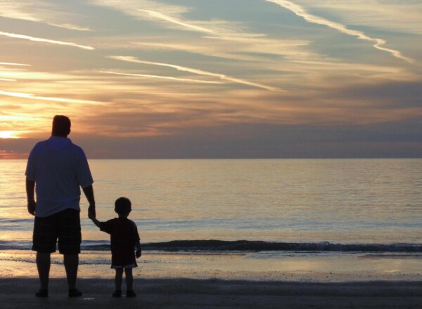 Silhouette of father and young son holding hands on beach, looking out to sea