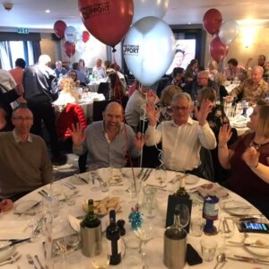 People sitting around dining table, waving to camera and celebrating with balloons