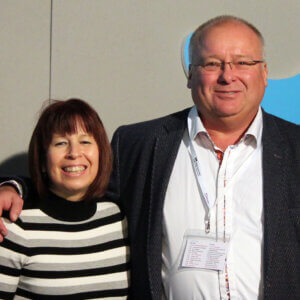 Man wearing lanyard standing next to woman, with arm around her shoulder, both smiling
