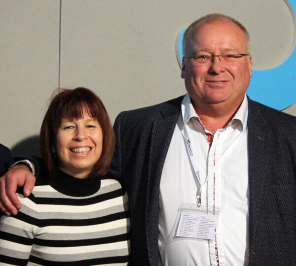 Man wearing lanyard standing next to woman, with arm around her shoulder, both smiling