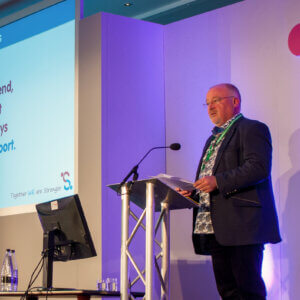 Man standing at lectern next to display screen, delivering a speech