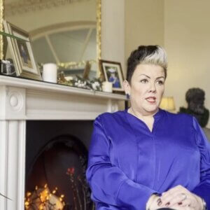 Image shows Ali sitting in a living room in front of a fireplace