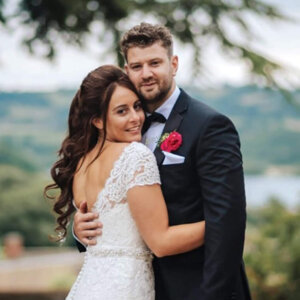 Bride with long dark hair, wearing lacy bridal dress, hugging her husband, both smiling