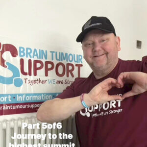 A man wearing a baseball cap and a Brain Tumour Support tshirt is standing next to a Brain Tumour Support charity banner and making the shape of a heart with his hands.