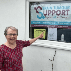 Angela by her front window pointing to fundraising information for Brain Tumour Support