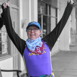 Angela in Brain Tumour Support bandana and a cap, smiling, arms raised triumphantly