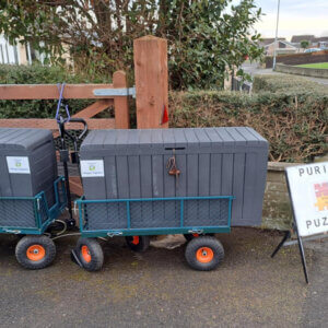 Two grey storgae boxes on wheels and a notice board standing at a driveway entrance