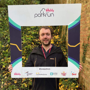 Young man standing by a wall holding a 'Parkrun' selfie frame, with trees and autumn leaves around