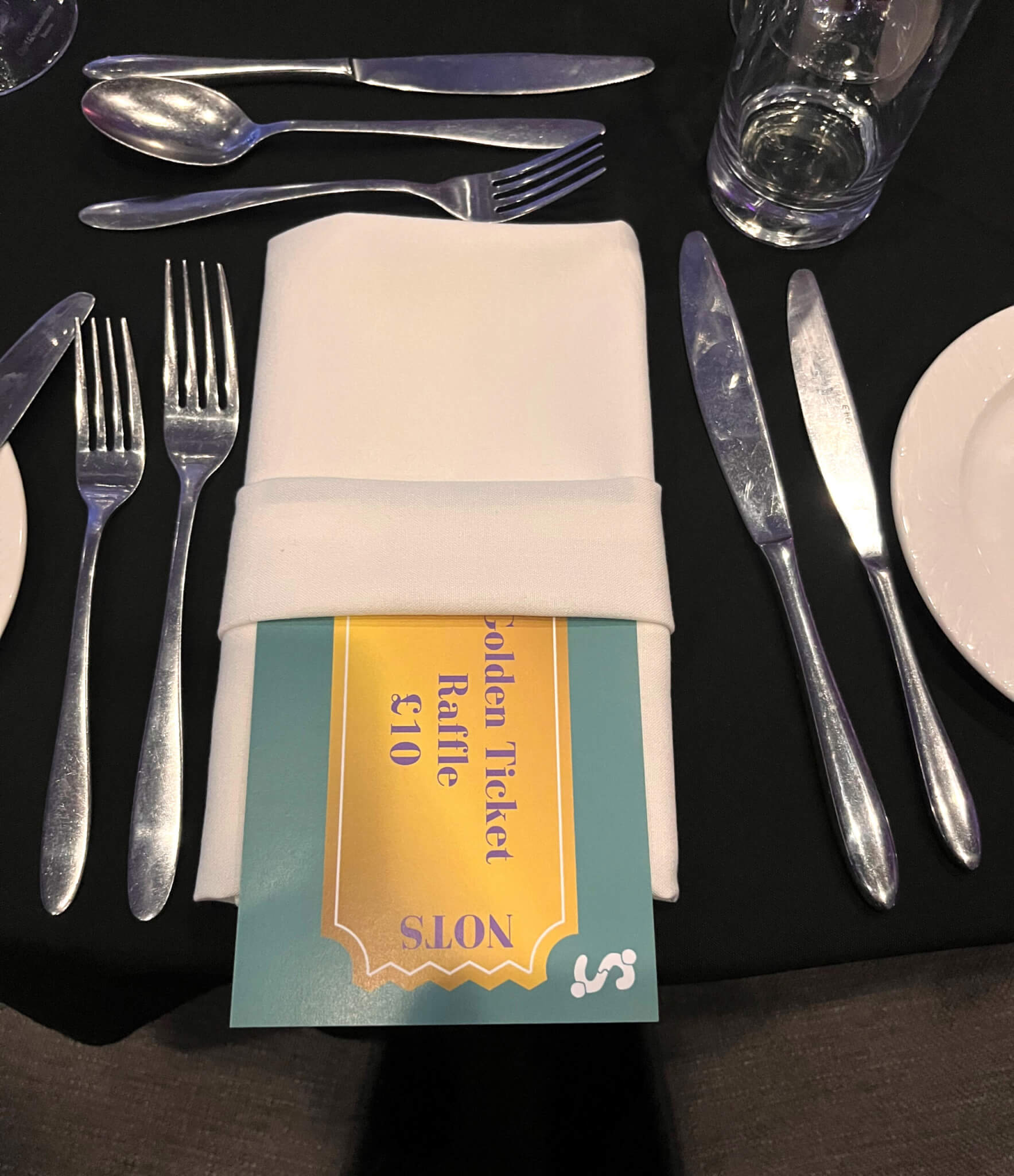 Large gold and green ticket tucked into a white napkin between smart silver cutlery