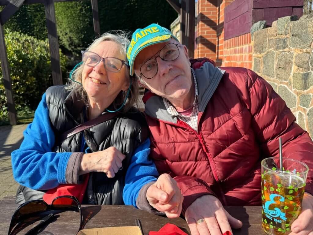 Older man in green cap and woman, both wearing glasses, side by side, heads together affectionately