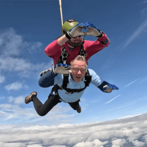 Two men above the clouds in tandem parachute jump - instructor holding hands up in a heart shape, elderly man below in Brain Tumour Support t-shirt with his hands out, smiling