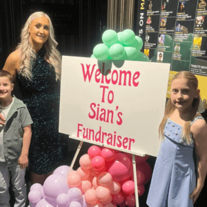 Sian fundraising for her Brain Tumour Support Champion Fund. At her event with two children and a sign covered in balloons.