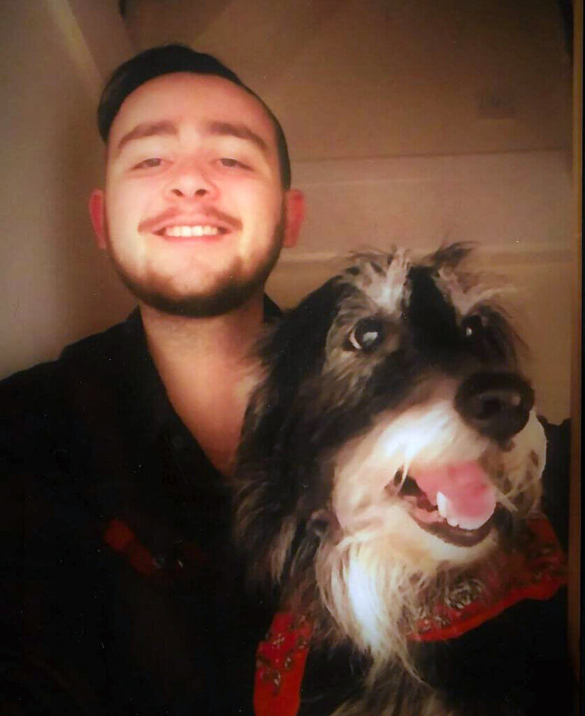 Young man with a neat beard, smiling, next to a happy looking long haired terrier dog