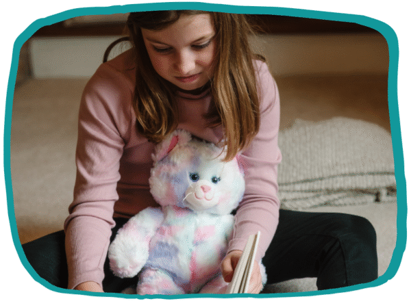 Illustration of Alice sitting on the floor with her soft teddy on her lap, reading a book and looking thoughtful, outlined with an irregular teal frame.