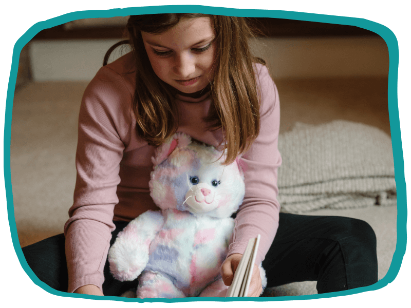 Illustration of Alice sitting on the floor with her soft teddy on her lap, reading a book and looking thoughtful, outlined with an irregular teal frame.