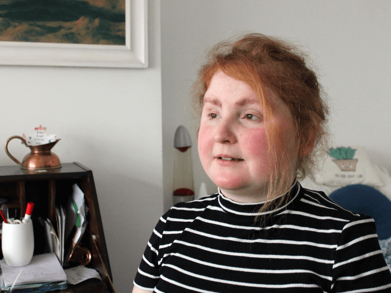 Young red-haired woman in black and white striped top, seated in bedroom, thoughtful and looking slightly away from camera
