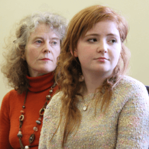 Mum sitting just behind red-haired late-teenage daughter, both thoughtful, looking off camera